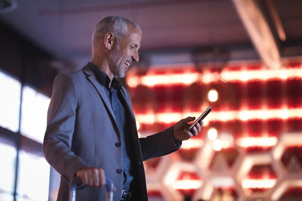A traveler in a blazer checks his phone while holding a suitcase handle in an airport terminal lit by red overhead panels.