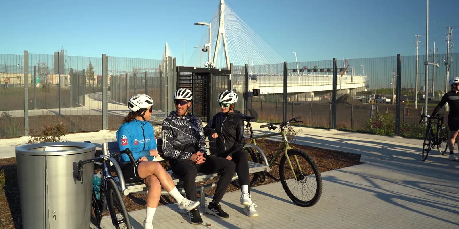 Cyclists resting on a bench at the trailhead of the Gordie Howe International Bridge multi-use path, with the bridge tower behind them