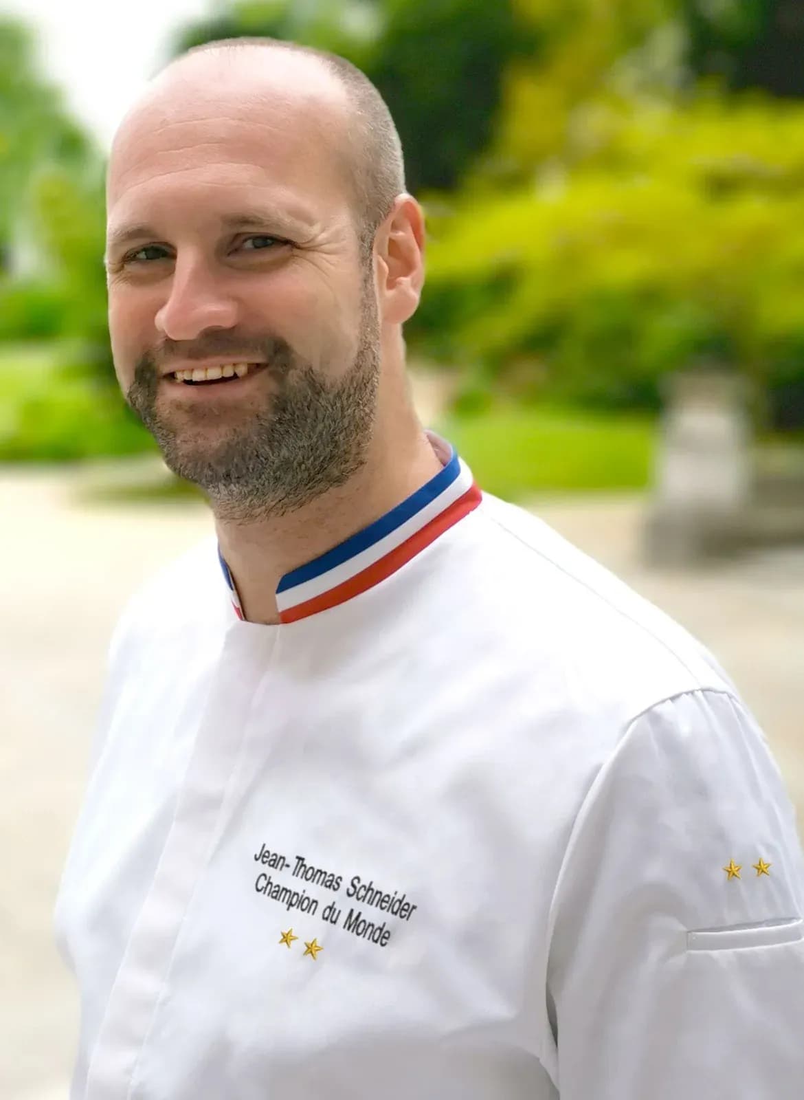 A bearded chef in a white jacket with a blue, white and red French tricolor collar stands in a sunlit garden, his name embroidered on the chest.
