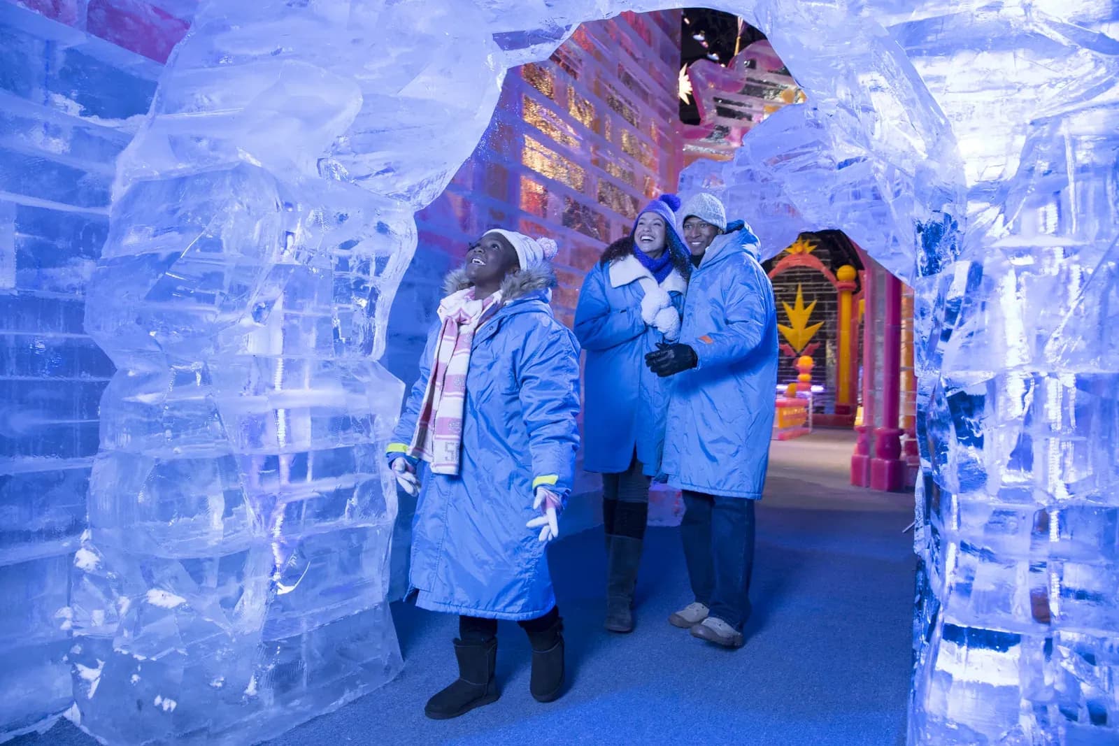 Visitors in blue parkas walk through a hand carved ice tunnel lit with colored lights at Gaylord Hotels ice! Christmas attraction