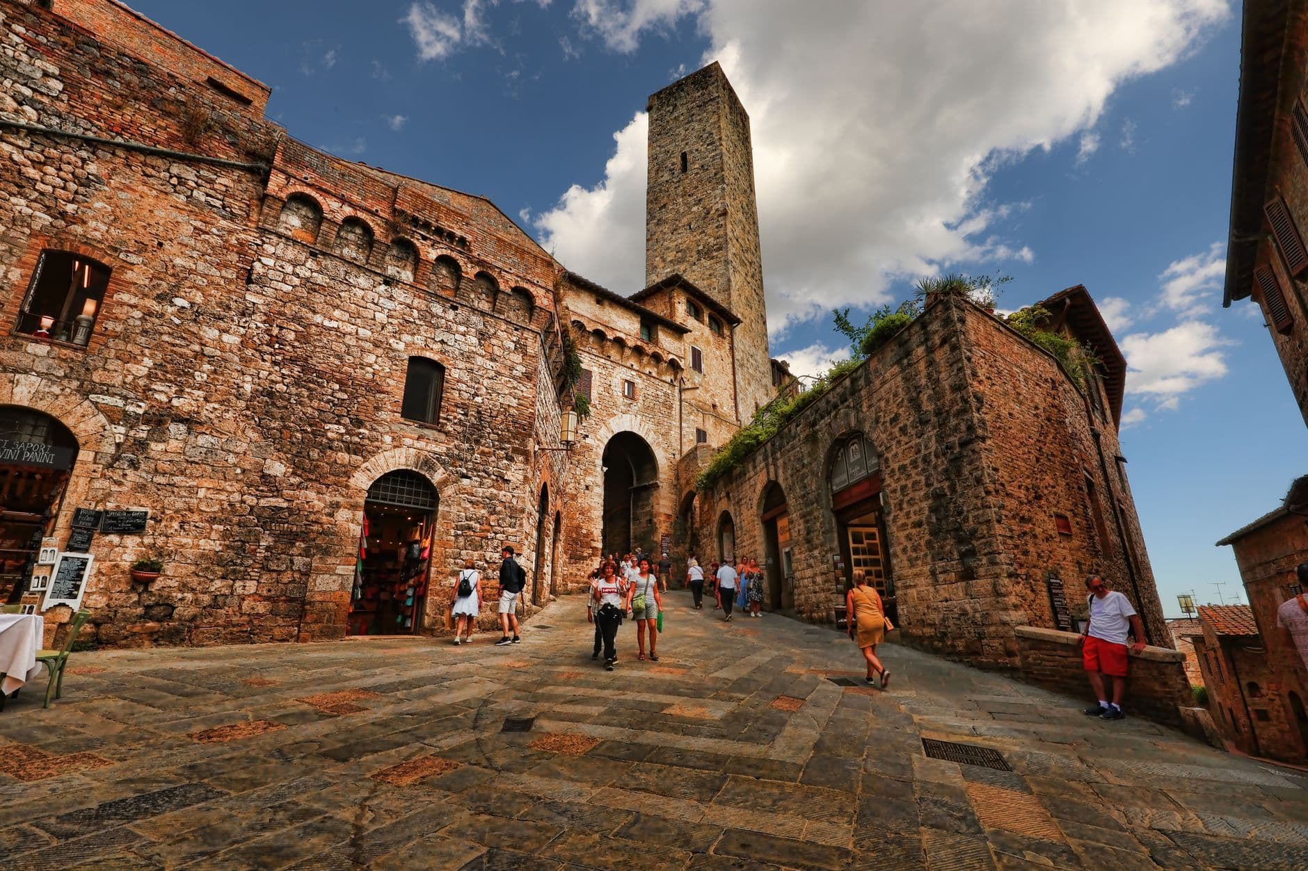 Travellers walk through a sunlit stone piazza beneath a medieval tower in a hilltop town.