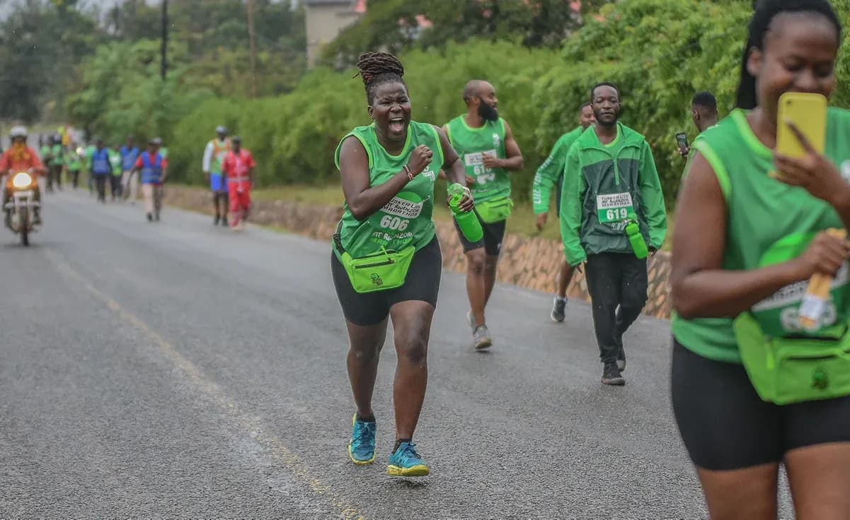 A woman wearing a green Tusker Lite Rwenzori Marathon vest and bib number 606 runs along a wet tarmac road in the hills near Kasese, with other runners and a motorcycle escort behind her.