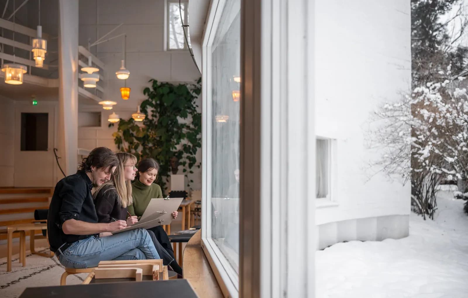 Three people sketch on a bench beside the curved window of Studio Aalto in Helsinki, with snow outside.